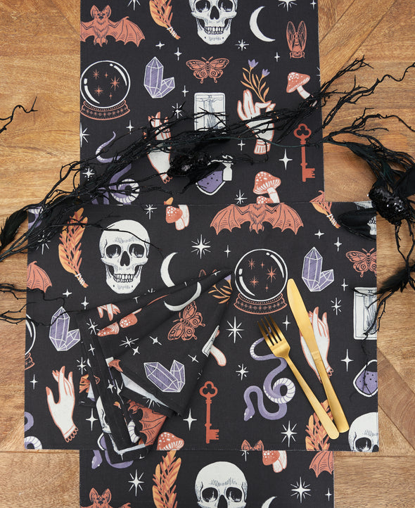 black table linens with mystical decorative elements including tarot cards, crystal ball, skulls, and other Halloween inspired motifs on a wood table