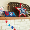 a hooked pillow shaped like a star with a red border and white dark blue and light blue stars that form smaller stars on a mantle next to a patriotic garland and additional americana gift pillows