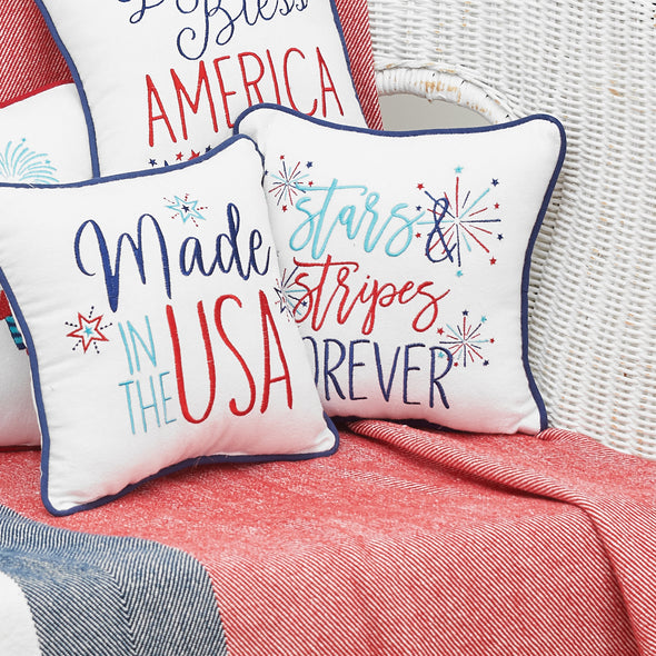 an embroidered pillow with blue and red flourishing text on a white background surrounded by bursts of red and blue fireworks and trimmed in a complementary blue surrounded by other patriotic gift pillows