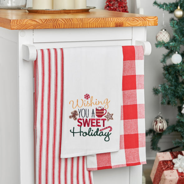 embroidered flour sack towel with the phrase wishing you a sweet holiday surrounded by classic Christmas sweets including a gingerbread man, peppermint candy, hot cocoa, and candy cane hanging from a kitchen countertop