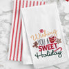 embroidered flour sack towel with the phrase wishing you a sweet holiday surrounded by classic Christmas sweets including a gingerbread man, peppermint candy, hot cocoa, and candy cane on a marble countertop