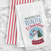 embroidered flour sack towel with the phrase Walking in a Winter Wonderland above a snow globe featuring a snowman in a winter scene next to a tree on a marble countertop