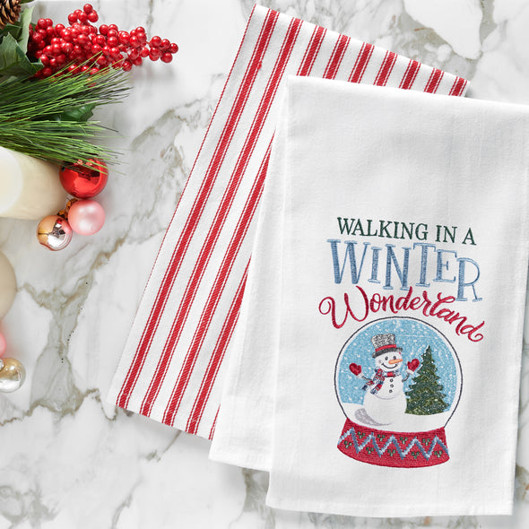 embroidered flour sack towel with the phrase Walking in a Winter Wonderland above a snow globe featuring a snowman in a winter scene next to a tree on a marble countertop