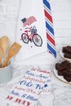 a patriotic waffle weave kitchen towel with a bike surrounded by stars and a waving flag hanging on a wall and surrounded by other kitchen decor